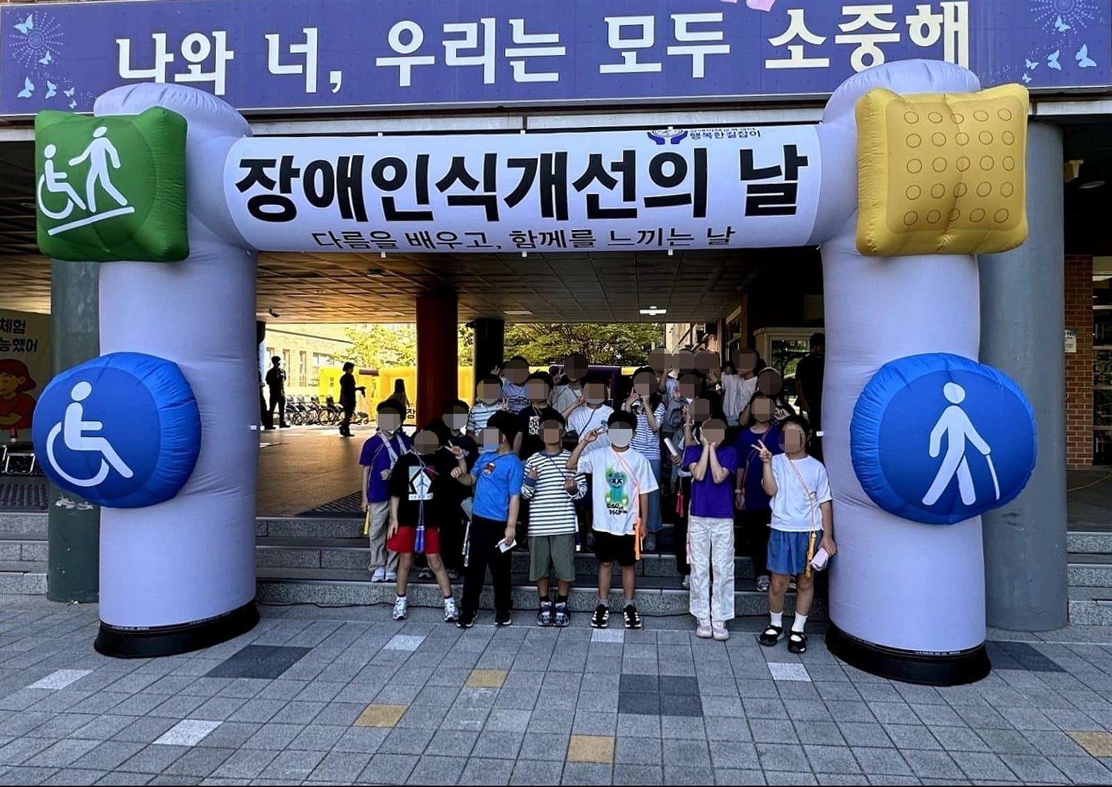 장애인식개선의 날 행사장 아치 아래 모인 초등학생들이 밝게 웃으며 브이 포즈를 하고 있다